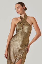 A woman is wearing a sparkling, metallic gold evening gown with a fitted silhouette. The dress features a halter neckline that crosses over at the neck and a small keyhole cutout in the front. It has a thigh-high slit on one side, revealing part of her leg. She accessorizes with large, round earrings.